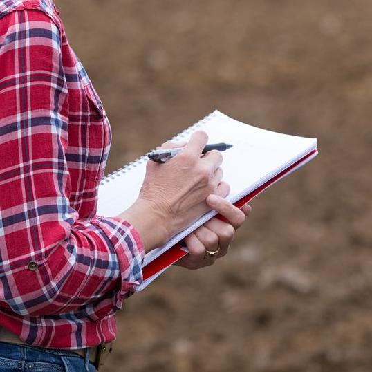 Zoom sur un carnet de prise de note tenu par une femme en plein champ cultivé
